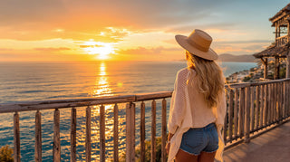 Woman in a hat and sunglasses standing on a wooden deck overlooking the ocean at sunset.