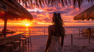 Woman standing on a beach at sunset, facing the ocean with straw umbrellas and tables in the foreground.