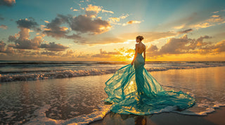 Woman in a flowing green dress standing on a beach at sunset.